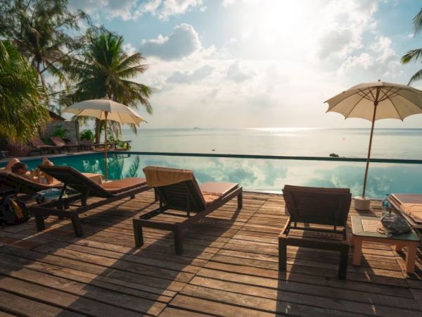 A serene poolside scene with lounge chairs and umbrellas on a wooden deck overlooking a calm body of water, surrounded by tropical trees.