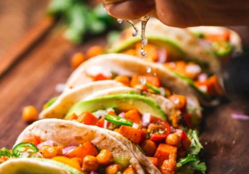 A hand is squeezing lime juice onto a row of tacos filled with chickpeas, avocado slices, cilantro, and diced vegetables arranged on a wooden board.