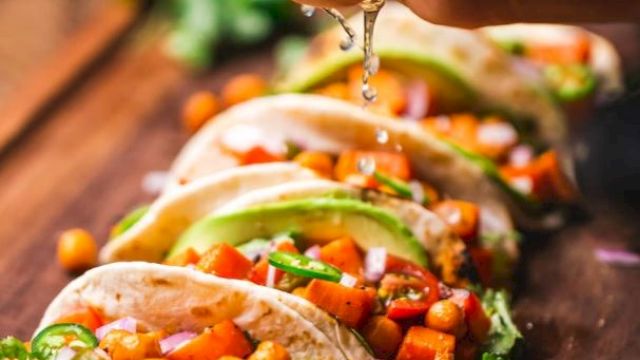 A hand is squeezing lime juice onto a row of tacos filled with chickpeas, avocado slices, cilantro, and diced vegetables arranged on a wooden board.