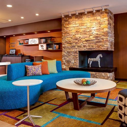 A modern, colorful lobby area with a blue curved sofa, round tables, a stone fireplace, and various decor elements, including a floor lamp and bookshelves.