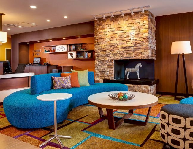 A modern, colorful lobby area with a blue curved sofa, round tables, a stone fireplace, and various decor elements, including a floor lamp and bookshelves.