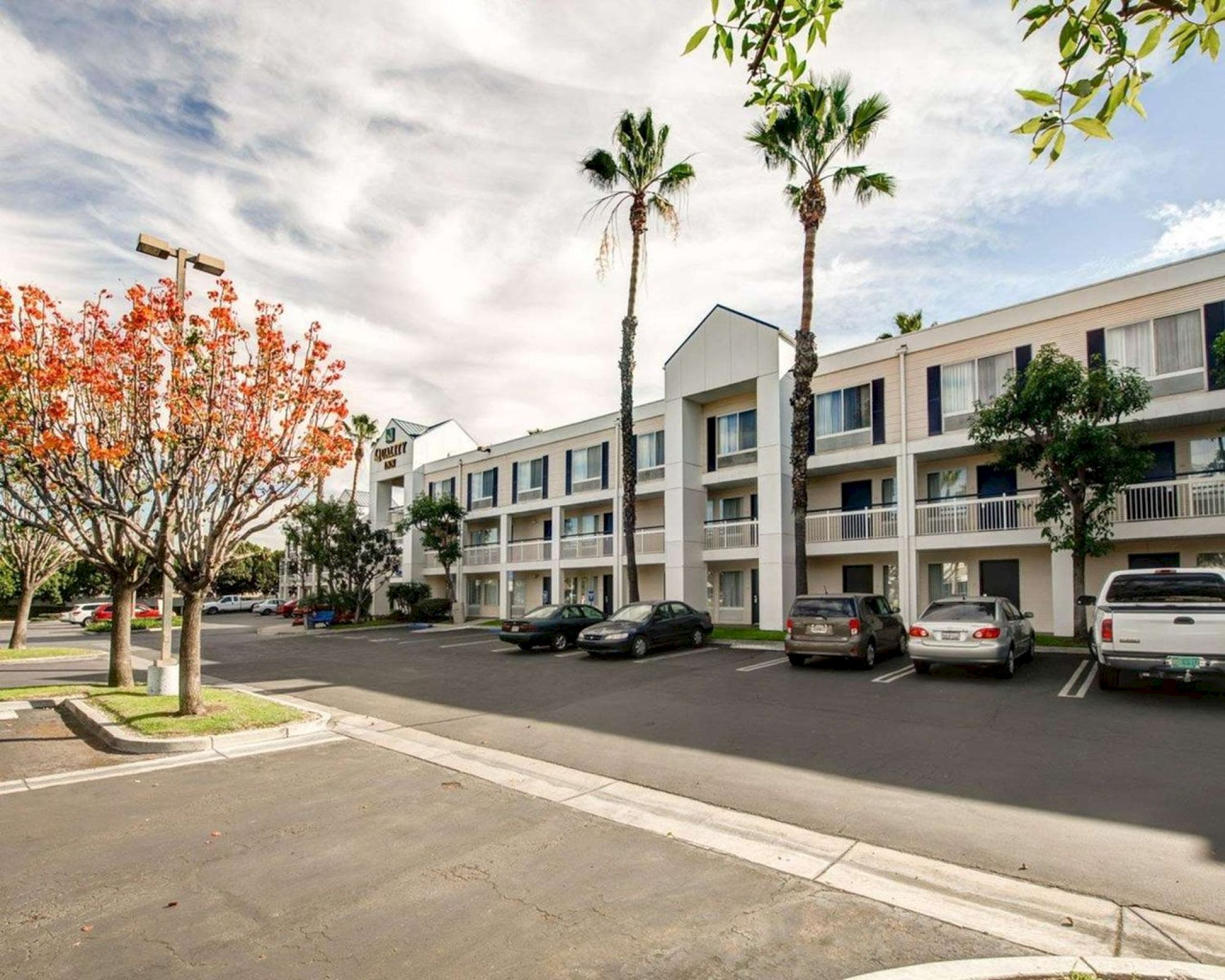 The image shows a three-story hotel or apartment building with a parking lot in front, several vehicles, and trees, including palm trees. The sky is partly cloudy.