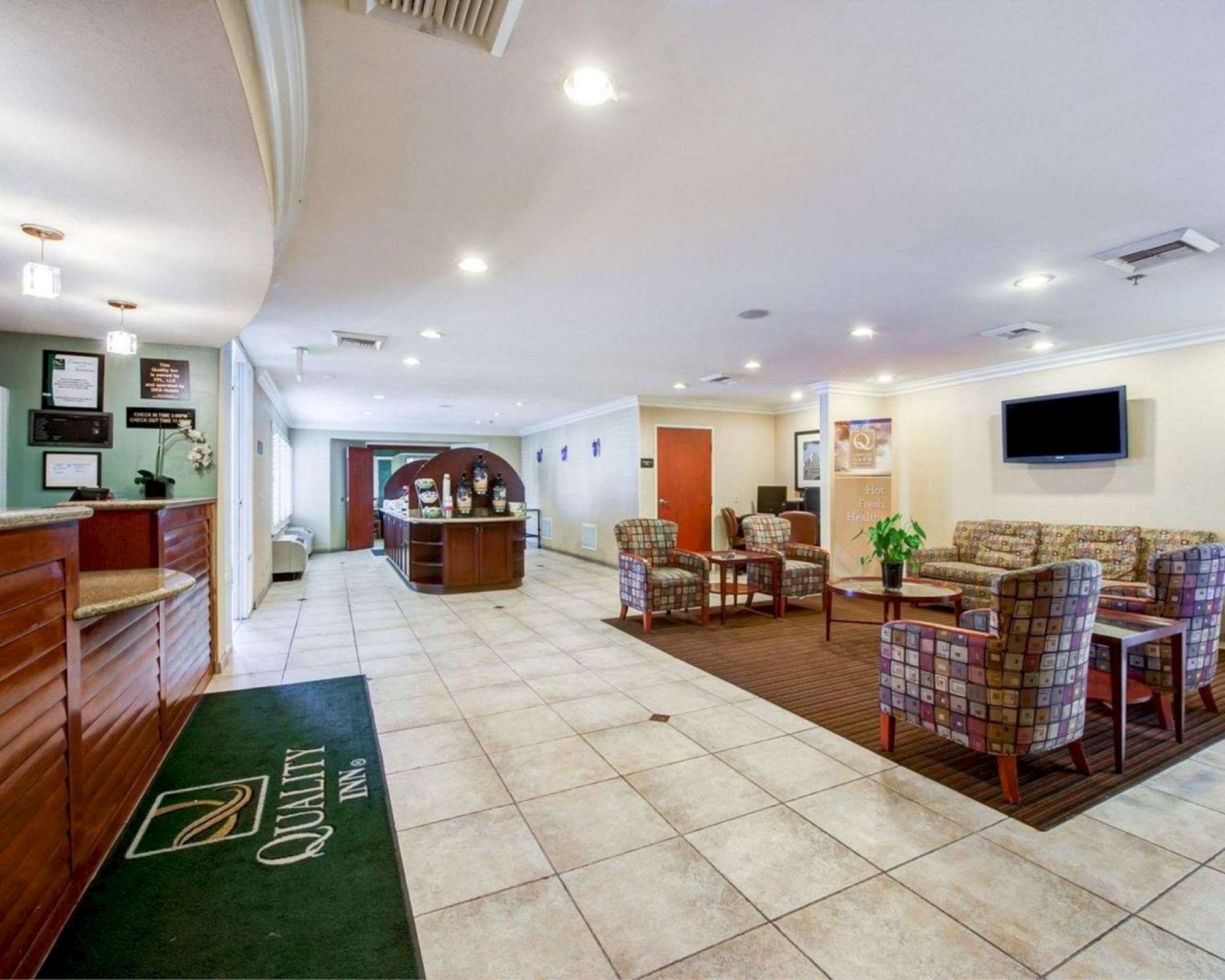 This image shows a hotel lobby with a tiled floor, a reception desk, seating area with armchairs and coffee tables, a TV, and decorative plants.