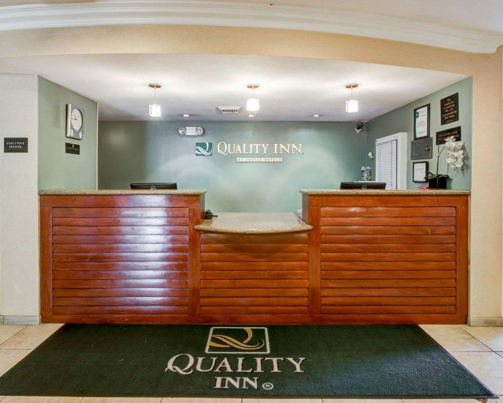 The image shows the reception desk of a Quality Inn hotel, featuring a green mat with the hotel’s logo and wooden paneling on the front desk.