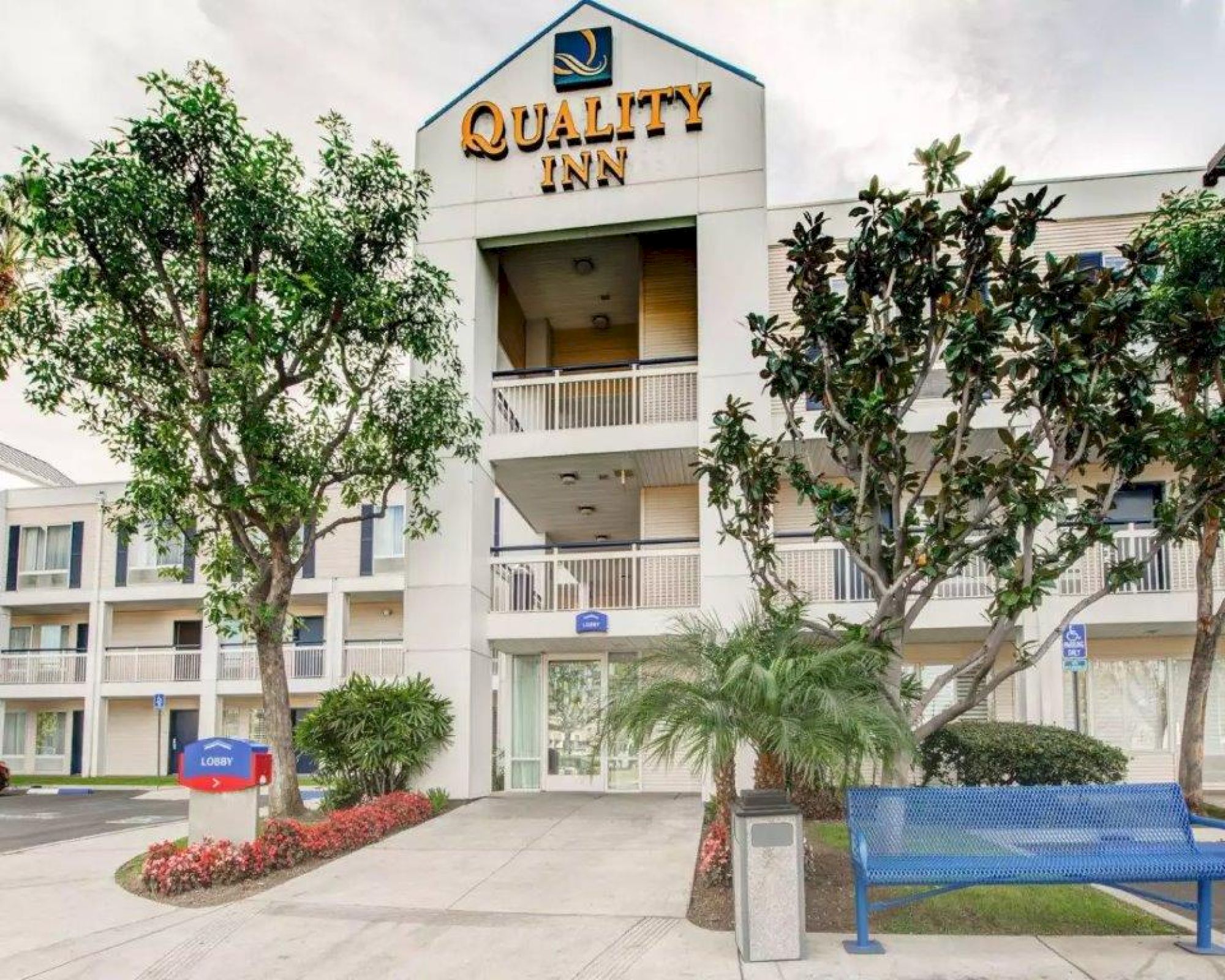 The image shows the exterior of a Quality Inn hotel, featuring a three-story building with balconies, surrounded by trees and landscaping.