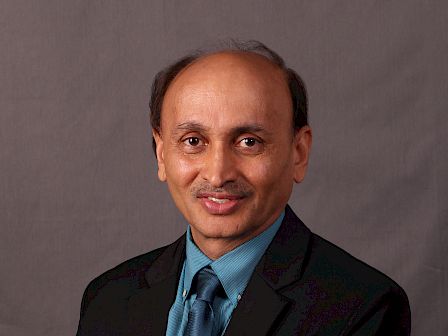 The image shows a man wearing a dark suit, blue shirt, and blue tie, standing against a grey background, smiling at the camera.