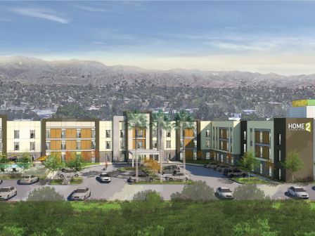 This image shows a hotel complex with a parking lot in front, situated against a backdrop of hills and a town in the distance.