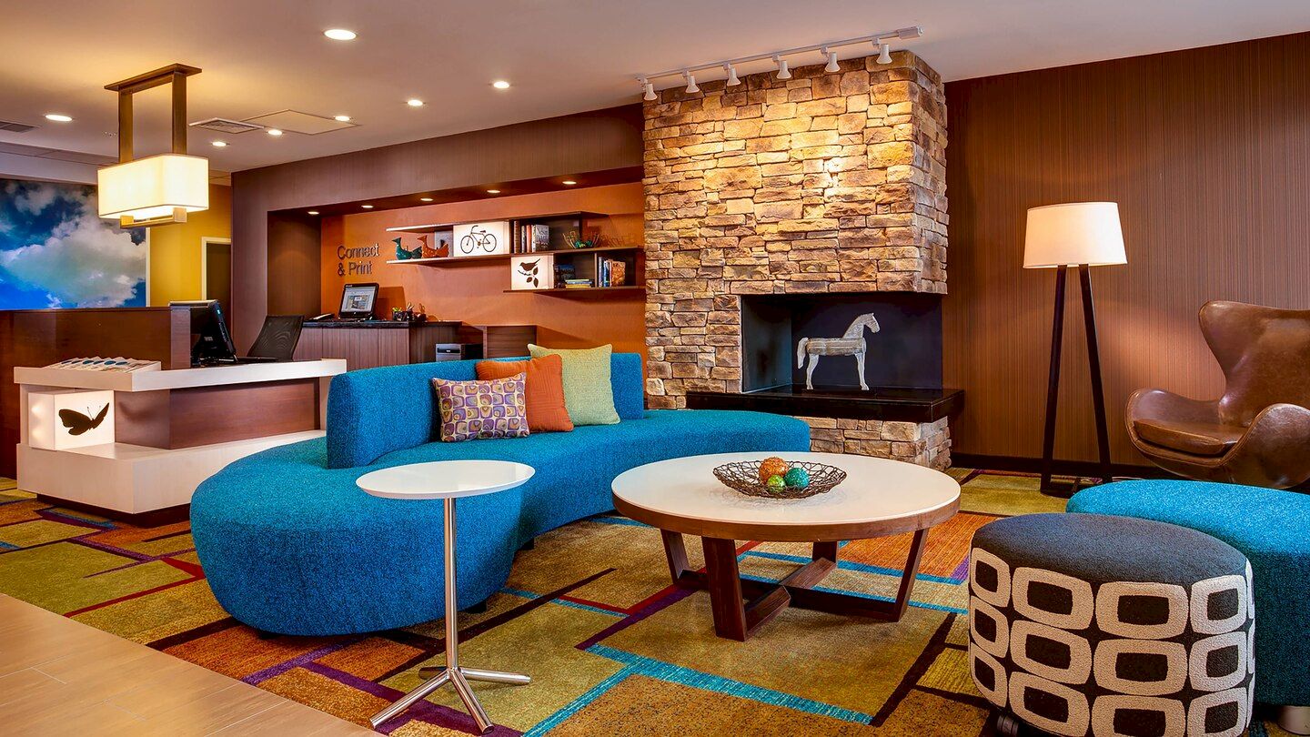 A modern hotel lobby with blue couches, a round coffee table, a stone fireplace, colorful carpet, a check-in desk, and contemporary decorations.