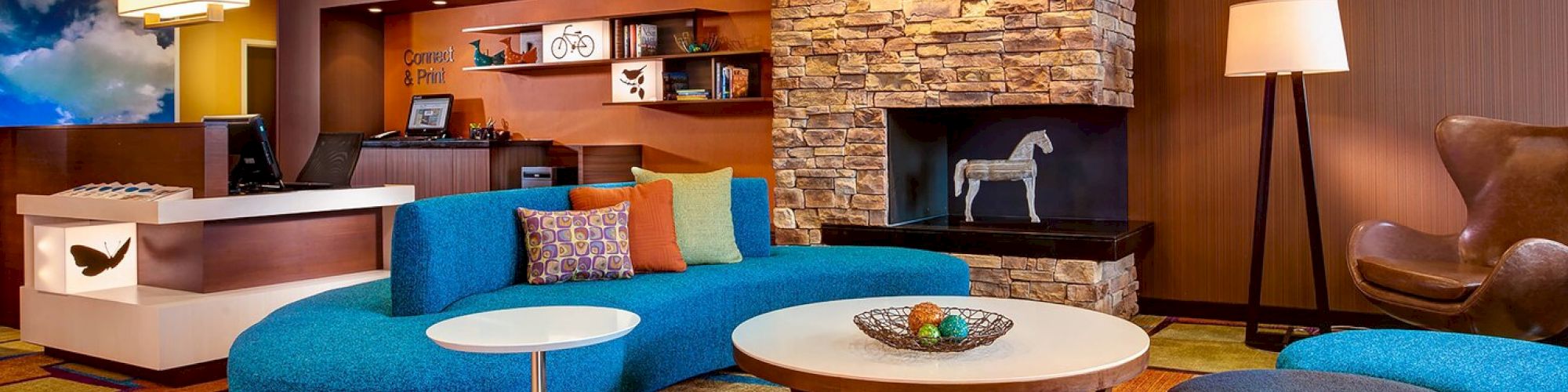 A modern hotel lobby with blue couches, a round coffee table, a stone fireplace, colorful carpet, a check-in desk, and contemporary decorations.