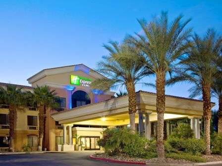 The image shows the exterior of a Holiday Inn Express & Suites hotel, featuring palm trees and a welcoming entrance, set against a blue sky.