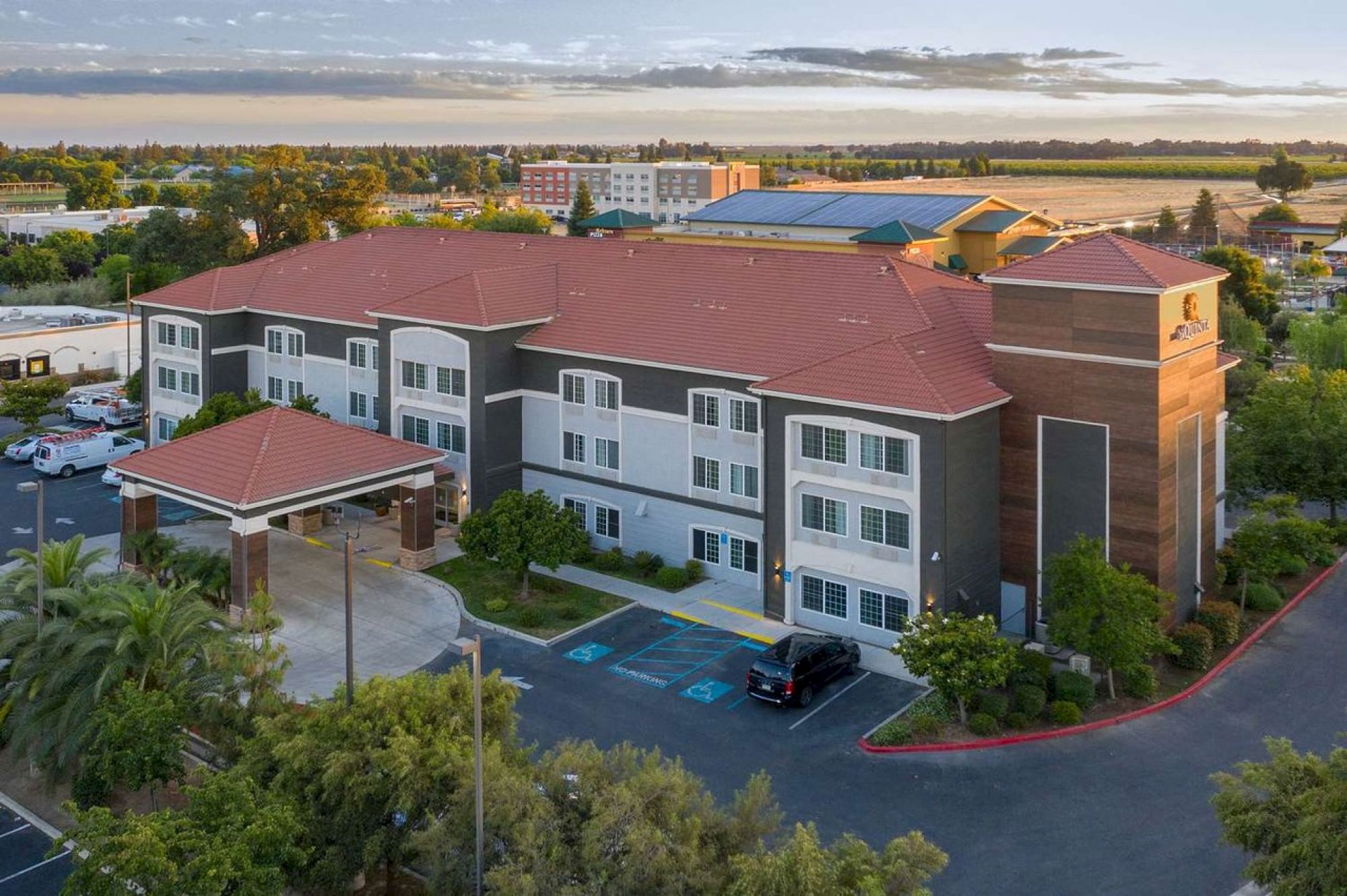 An aerial view of a three-story building with a red roof, a parking area, and greenery. The surroundings include more buildings and open fields.