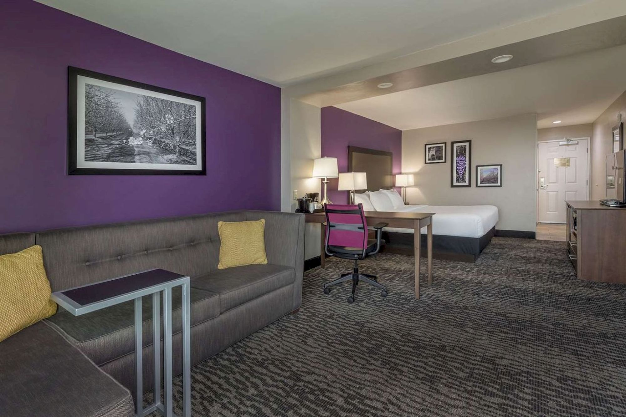 The image shows a modern hotel room with a bed, a couch, a desk and chair, and various wall art. The decor includes purple accents and neutral tones.