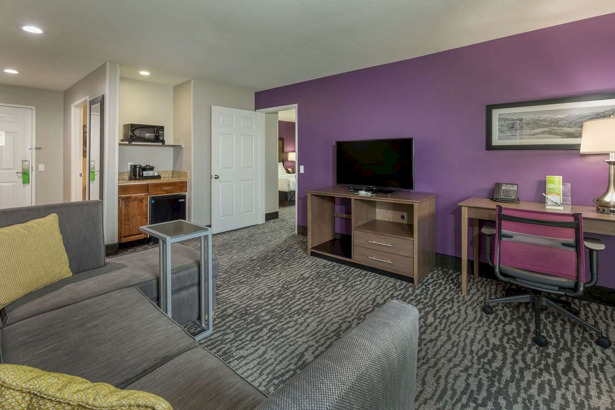 A cozy hotel room with a purple accent wall, sofa, TV, desk, kitchenette, and a bright lamp. The open door reveals a glimpse of a bed.