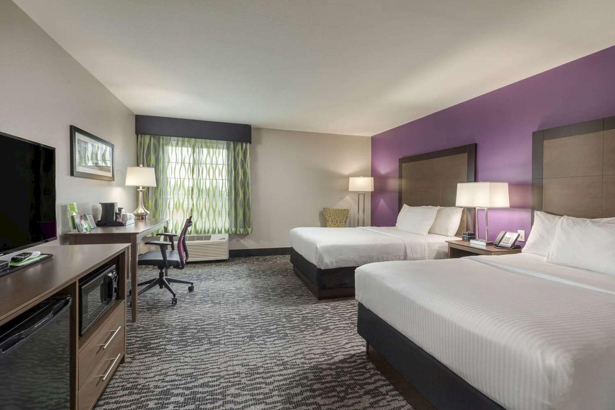 This image shows a modern hotel room with two double beds, a TV, a desk with a chair, and lamps on nightstands, with a purple accent wall.