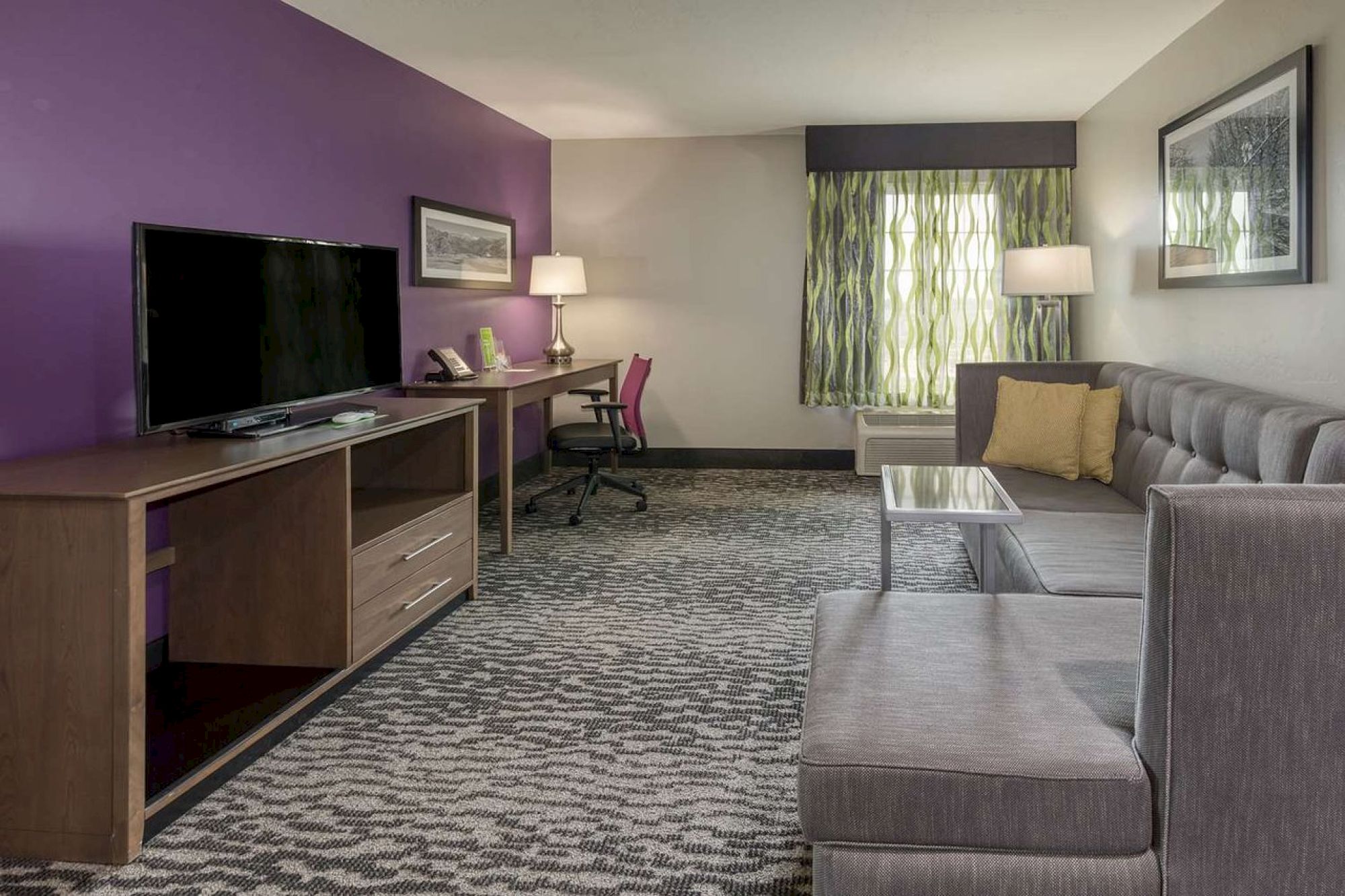 A modern hotel room with a purple wall, flat-screen TV, desk with chair, sofa, coffee table, floor lamp, and curtained window.