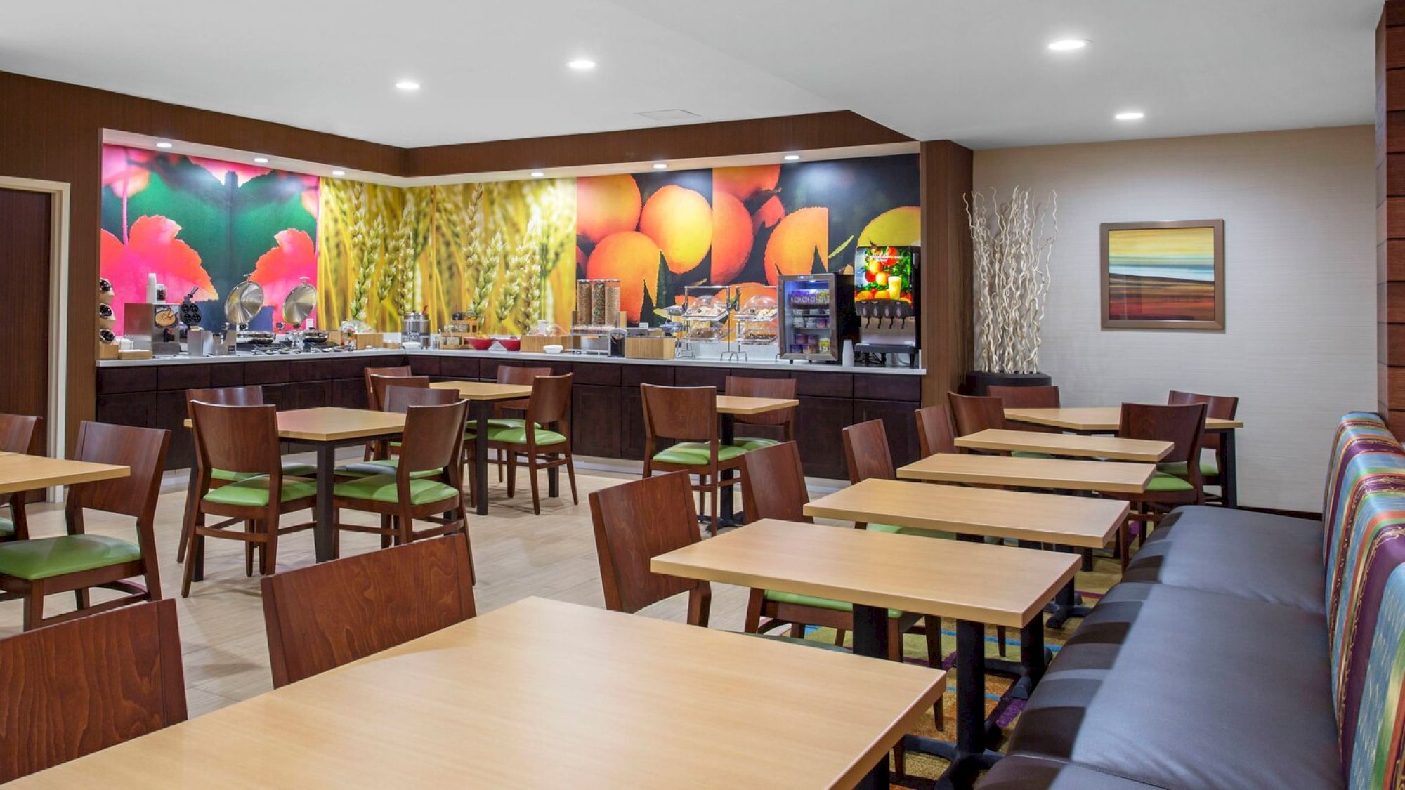 A modern dining area with tables and chairs, a colorful mural, and a breakfast buffet setup with various food items and beverages.