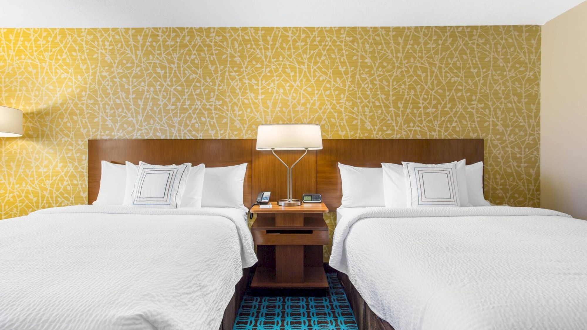 The image shows a hotel room with two neatly-made beds, a nightstand with a lamp and phone, and a patterned yellow wallpaper.