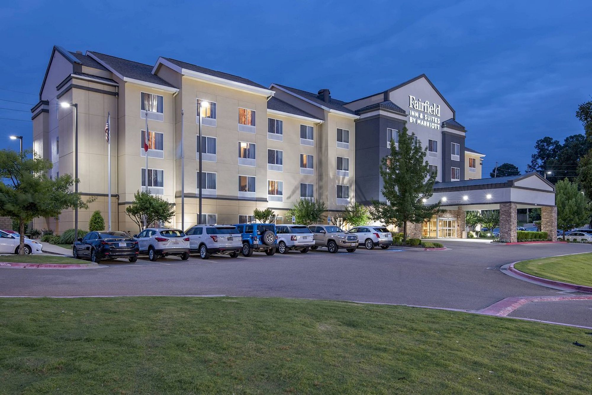 The image shows a four-story hotel building at dusk, with several cars parked in front, and trees and grass in the surrounding area.