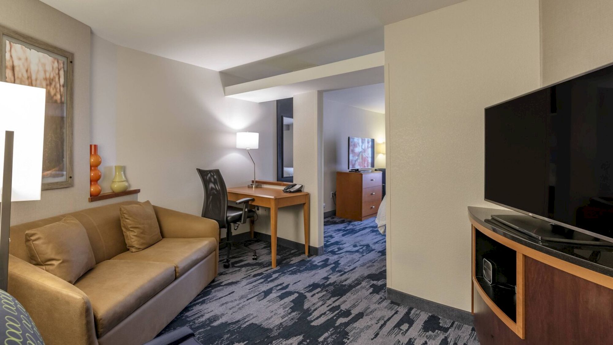 This image shows a cozy hotel room with a sofa, desk, chair, TV, and modern decor. The room has a carpeted floor and a neutral color palette.