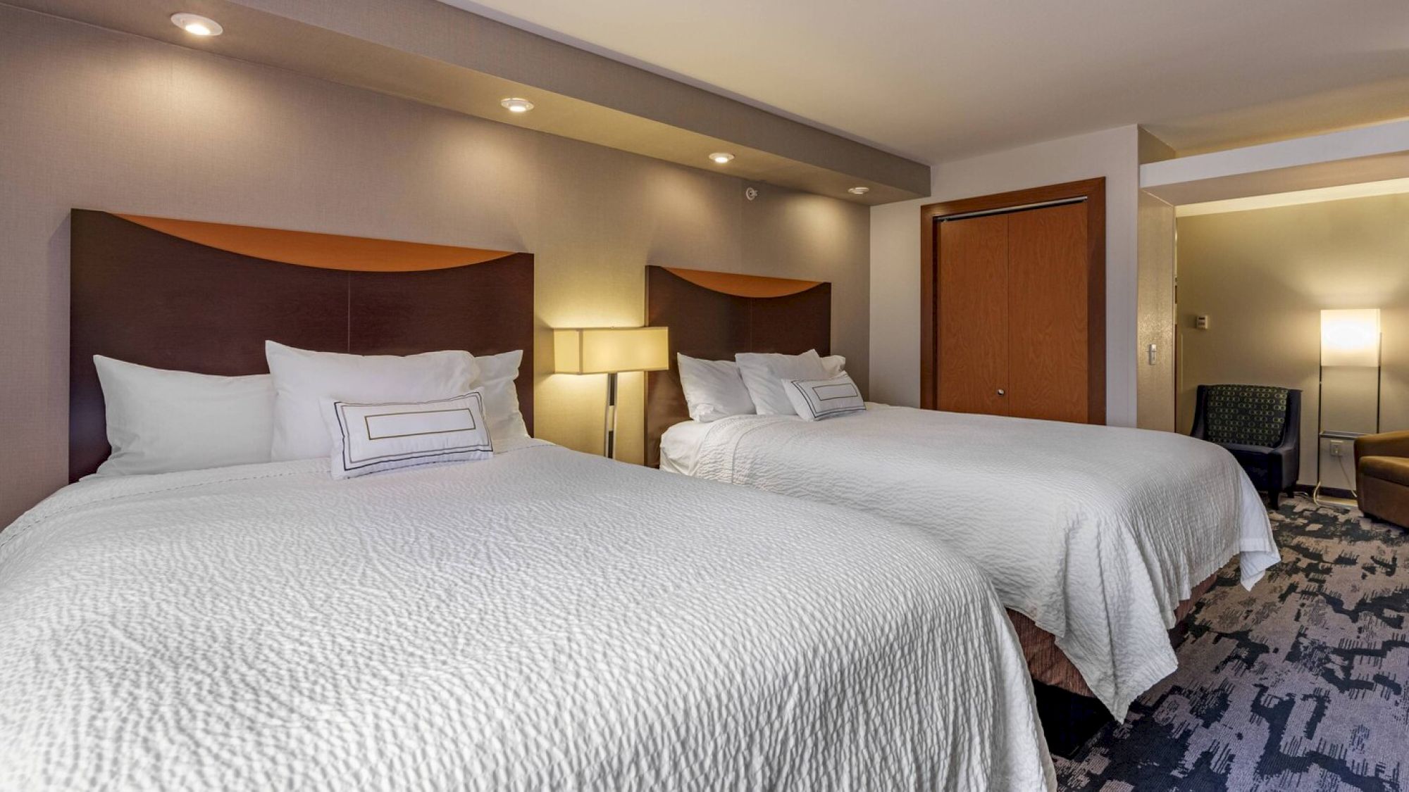 The image shows a hotel room with two neatly made queen-sized beds, a nightstand with a lamp between them, and modern decor.