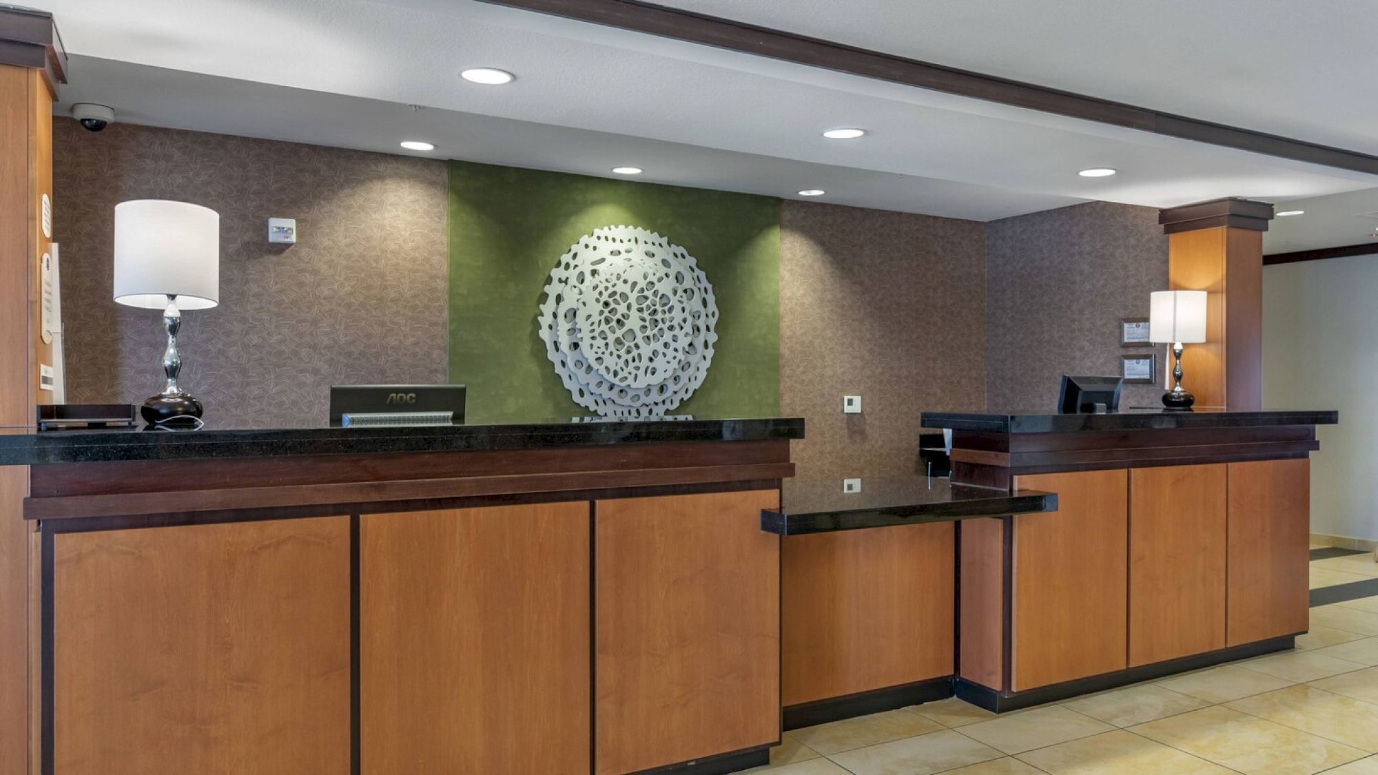 The image shows a hotel reception desk with wooden panels, a decorative wall piece behind, two lamps, and two computer stations for staff use.