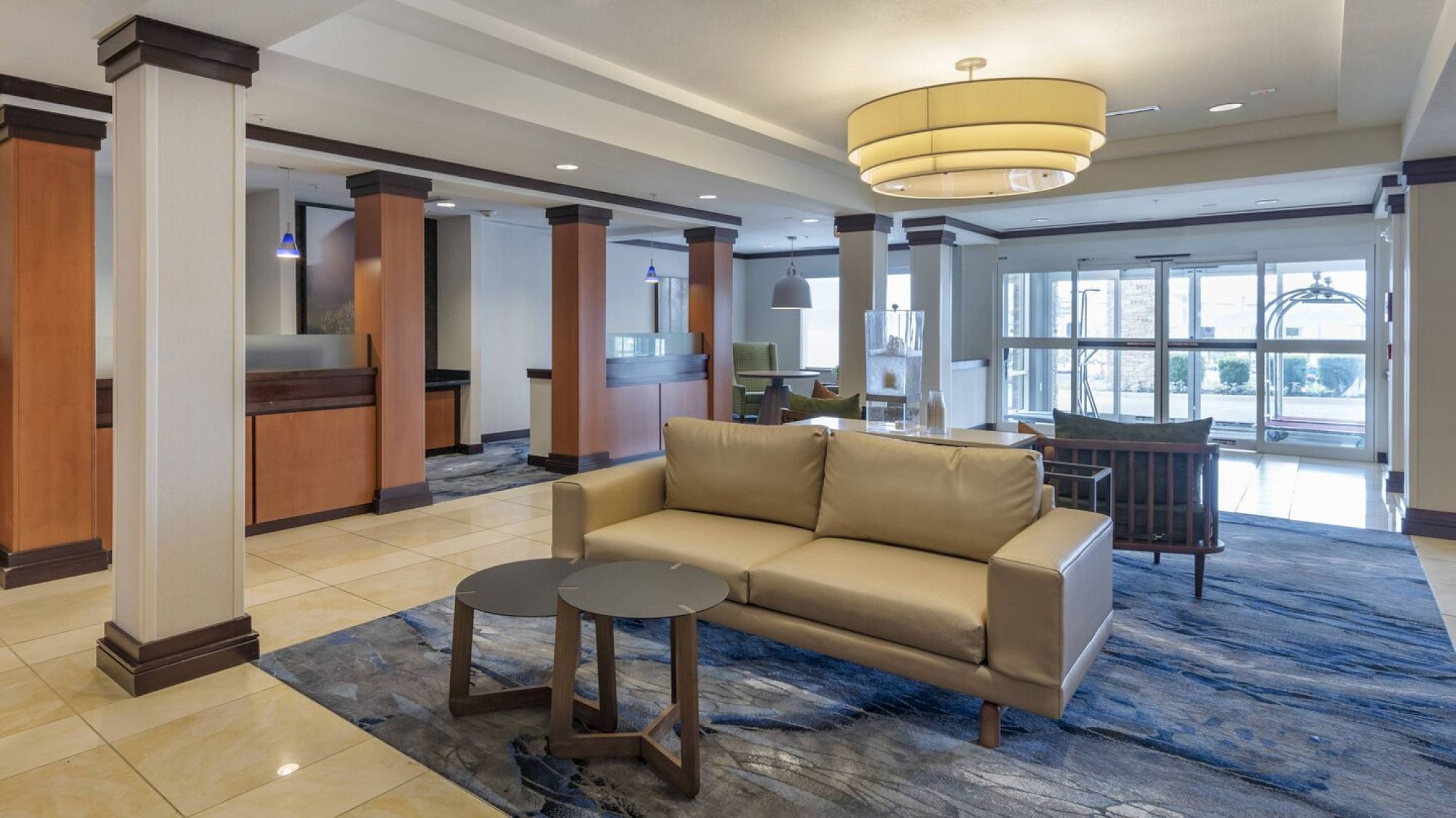 The image shows a modern hotel lobby with a beige sofa, small tables, a large ceiling light, and a reception desk in the background.