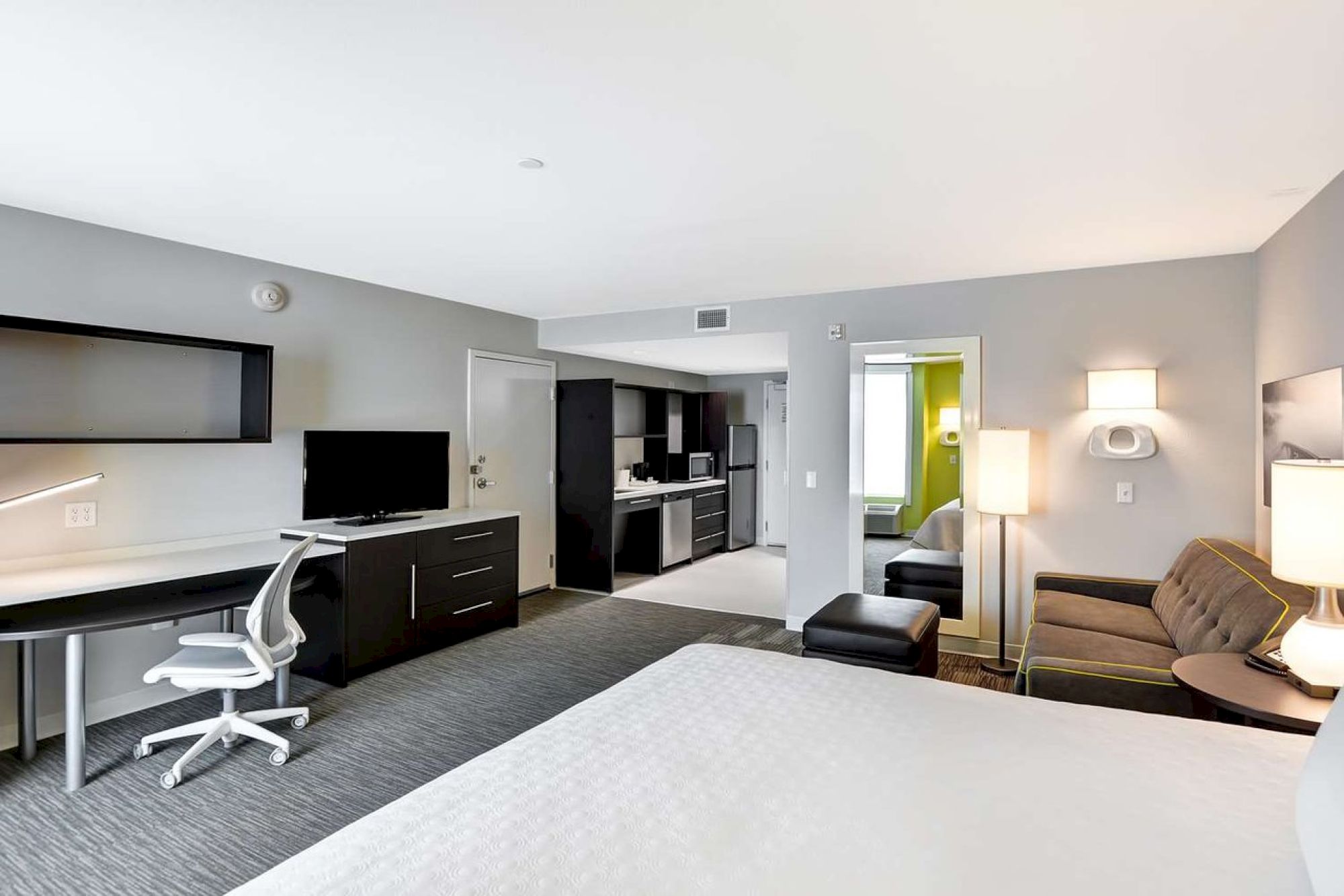 A modern hotel room with a bed, desk, TV, sofa, lamp, and a small kitchenette in the background, featuring neutral colors and minimalist decor.