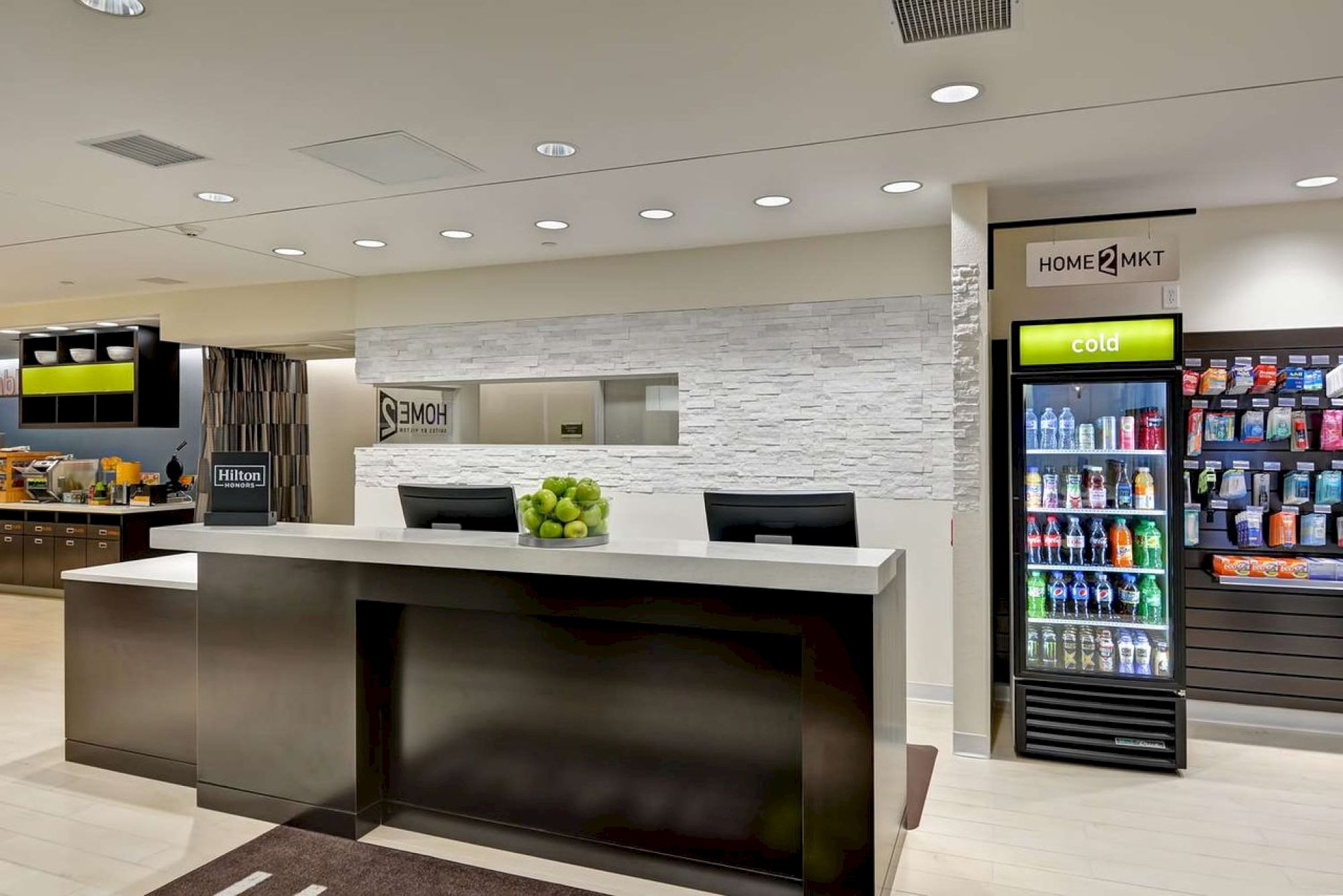 This image shows a modern hotel reception with two black counters, a bowl of green apples, a drink/snack vending machine, and a sign that reads "Home2."