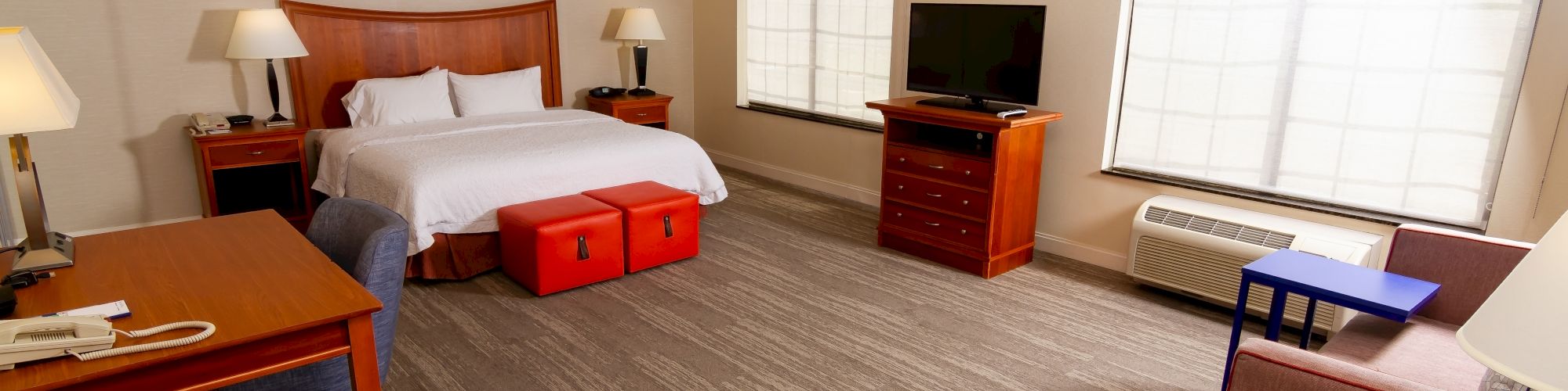 This image shows a hotel room with a bed, nightstands, desk, TV, sofa, and large windows with blue patterned curtains.