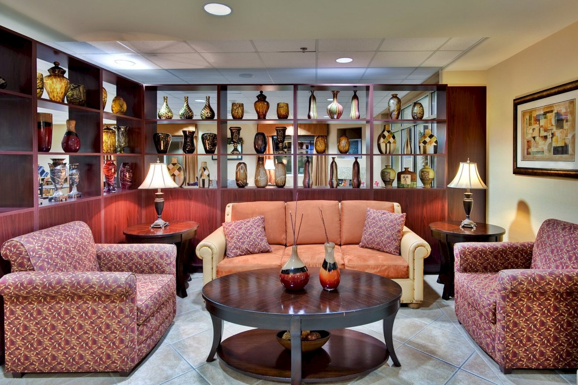 A cozy seating area with a sofa, two armchairs, a round coffee table, and shelves displaying vases and decorative items, plus three table lamps.