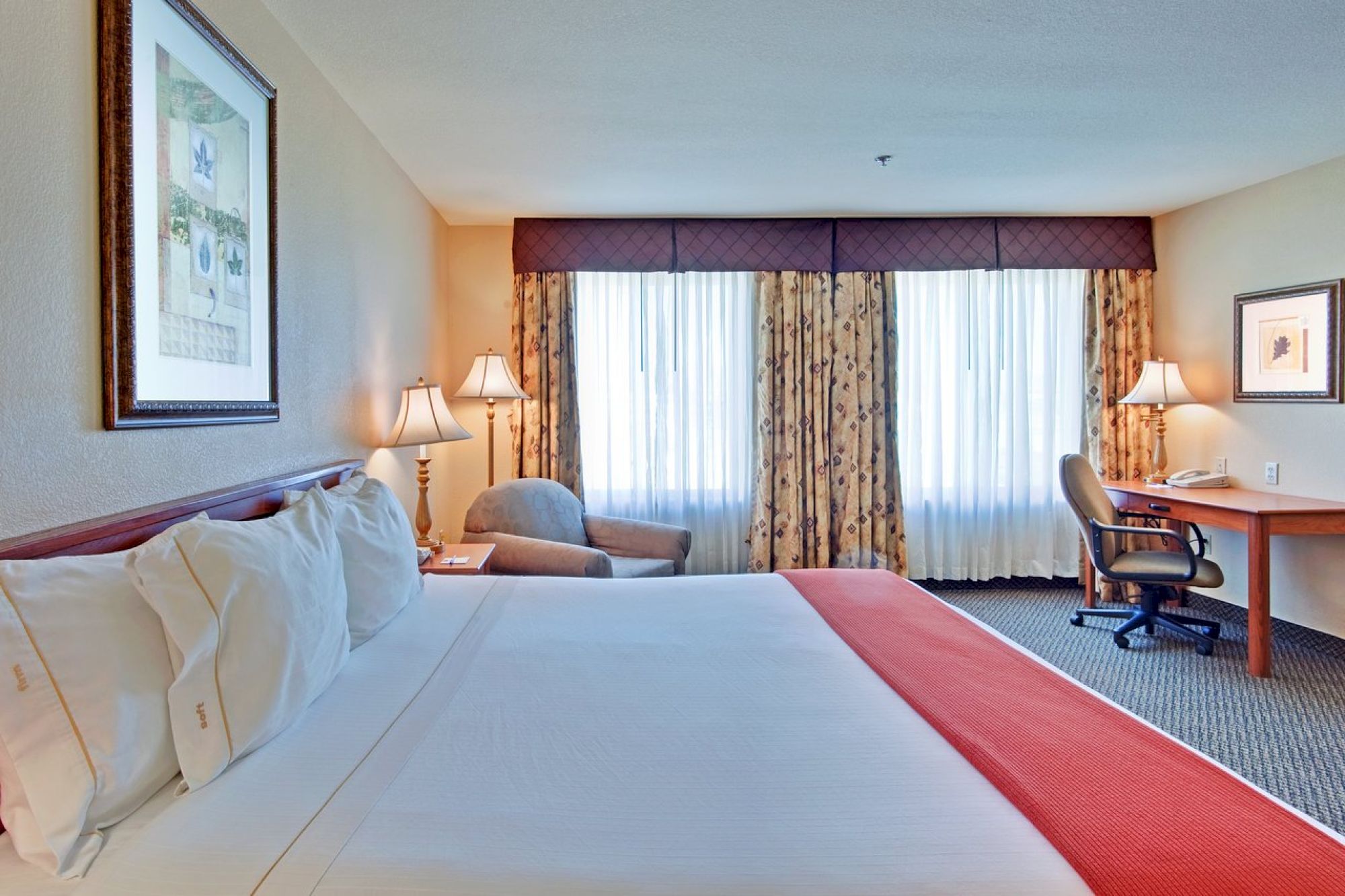 A hotel room with a neatly made bed, bedside lamps, armchair, desk with chair, windows with curtains, and framed artwork on the walls.