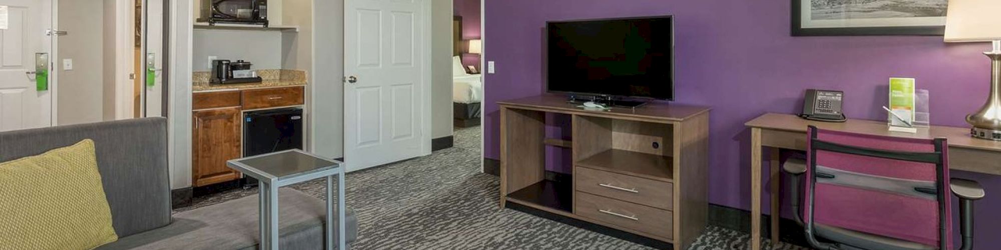 The image shows a modern hotel room with a gray sofa, purple accent wall, flat-screen TV, desk, and kitchenette area with a microwave and mini-fridge.