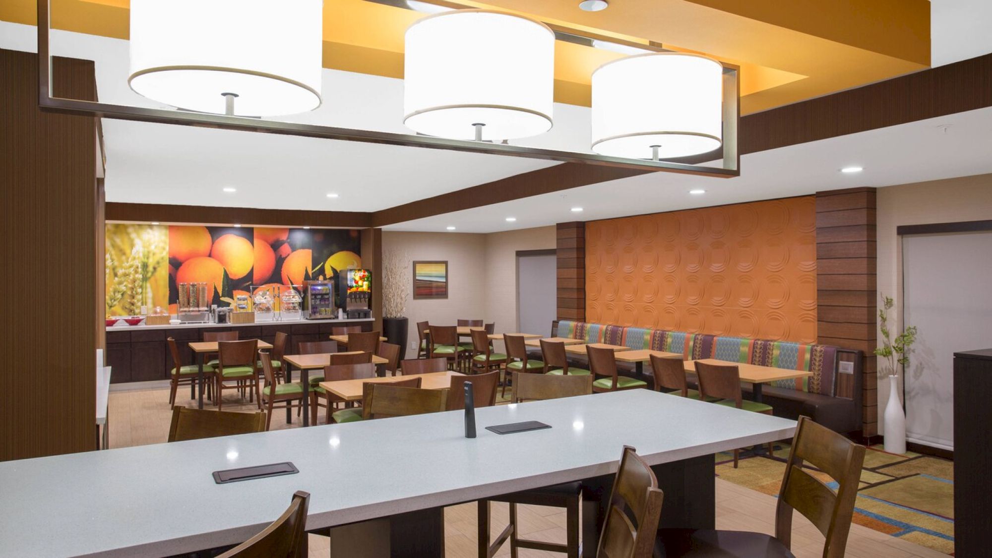 The image shows a modern, well-lit dining area with tables, chairs, a breakfast buffet, and contemporary decor, including vibrant wall art and lighting fixtures.