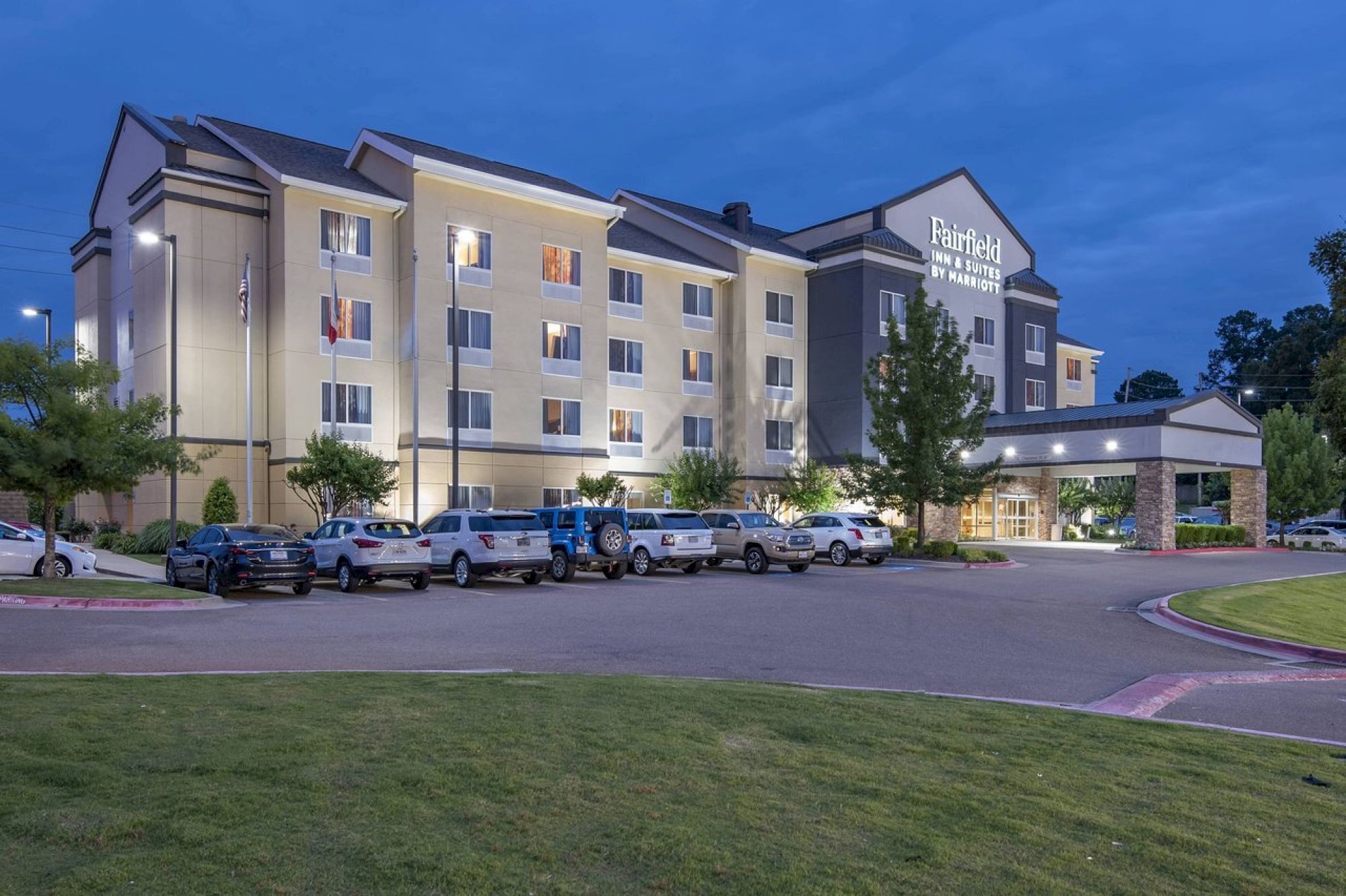 This image shows a multi-story hotel building with a parking lot in front, occupied by vehicles, and surrounded by landscaped greenery.