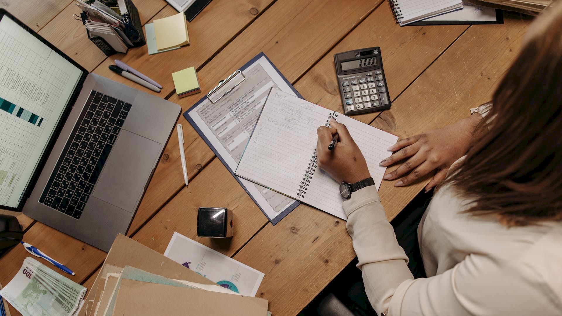 A person is working at a desk with a laptop, papers, calculator, and stationery, writing in a notebook while sitting in an office setting.