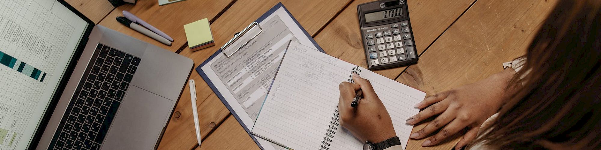 A person is working at a desk with a laptop, papers, calculator, and stationery, writing in a notebook while sitting in an office setting.