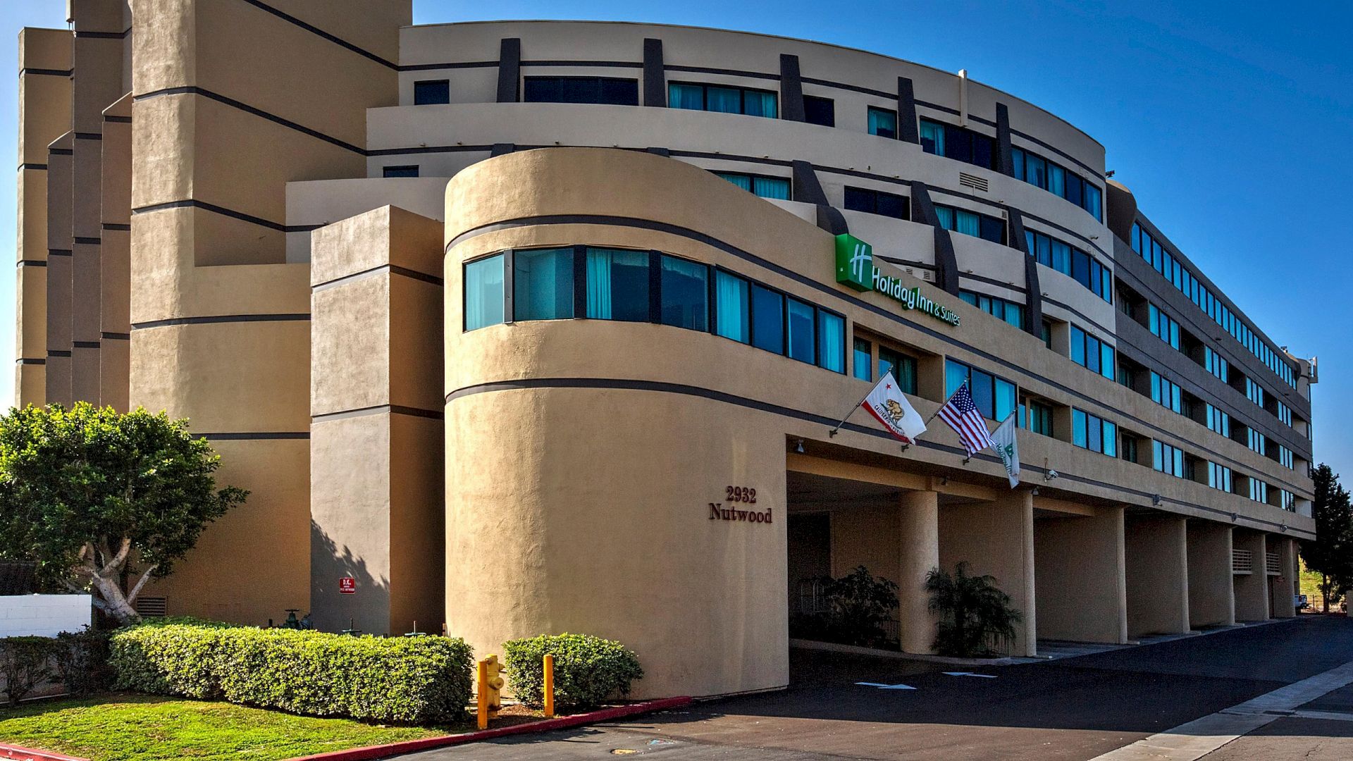 The image shows a modern, multi-story hotel building with a beige facade and green glass windows. Three flags are displayed at the entrance.