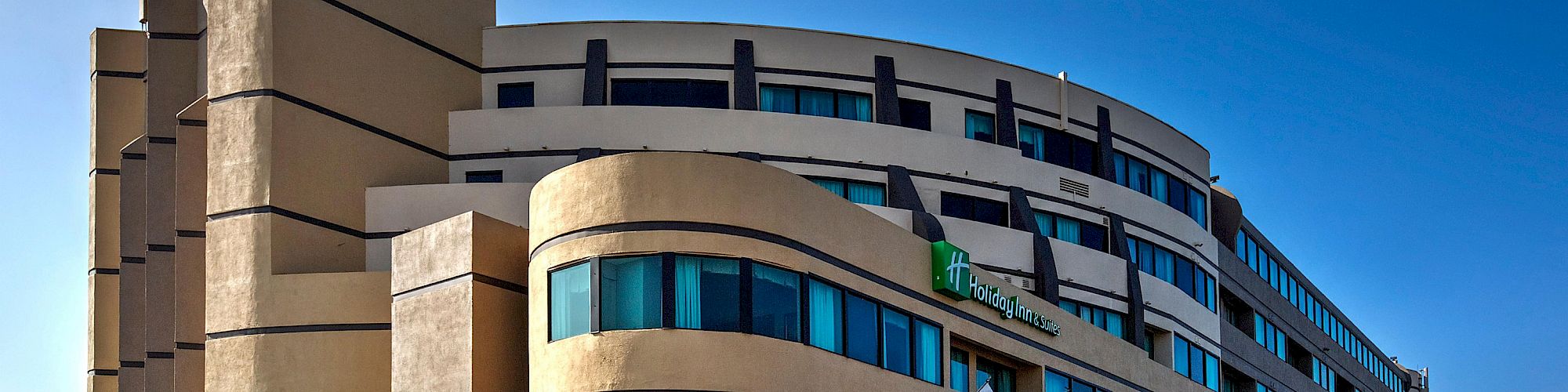 The image shows a modern, multi-story hotel building with a beige facade and green glass windows. Three flags are displayed at the entrance.