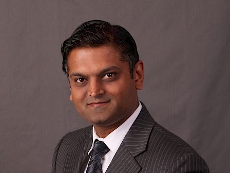 A person dressed in a dark pinstripe suit, white shirt, and patterned tie, posing against a plain gray background.