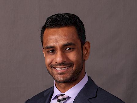 The image shows a man in a suit and tie smiling against a plain background.