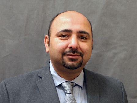 The image shows a man in a grey suit with a patterned tie, standing against a plain grey background.