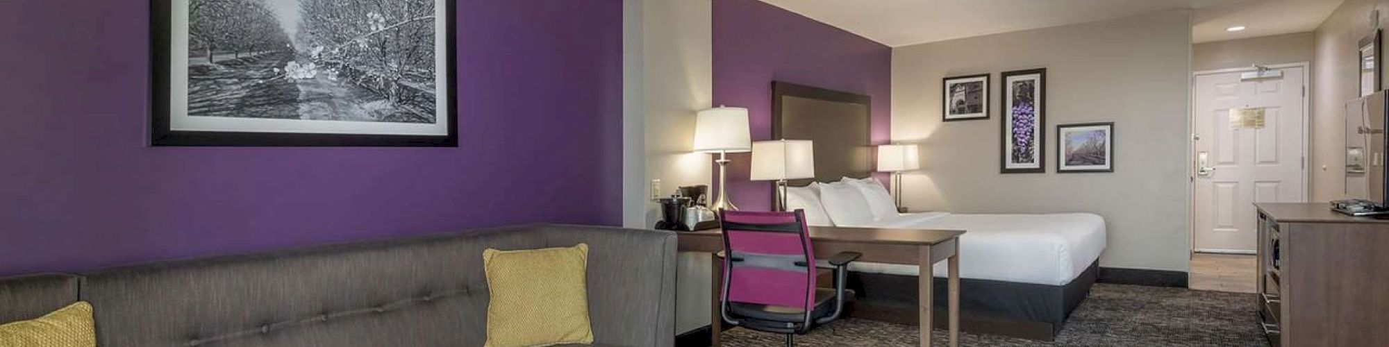 A modern hotel room with a purple accent wall, sofa, bed, desk, and lamps, decorated with framed pictures ending the sentence.