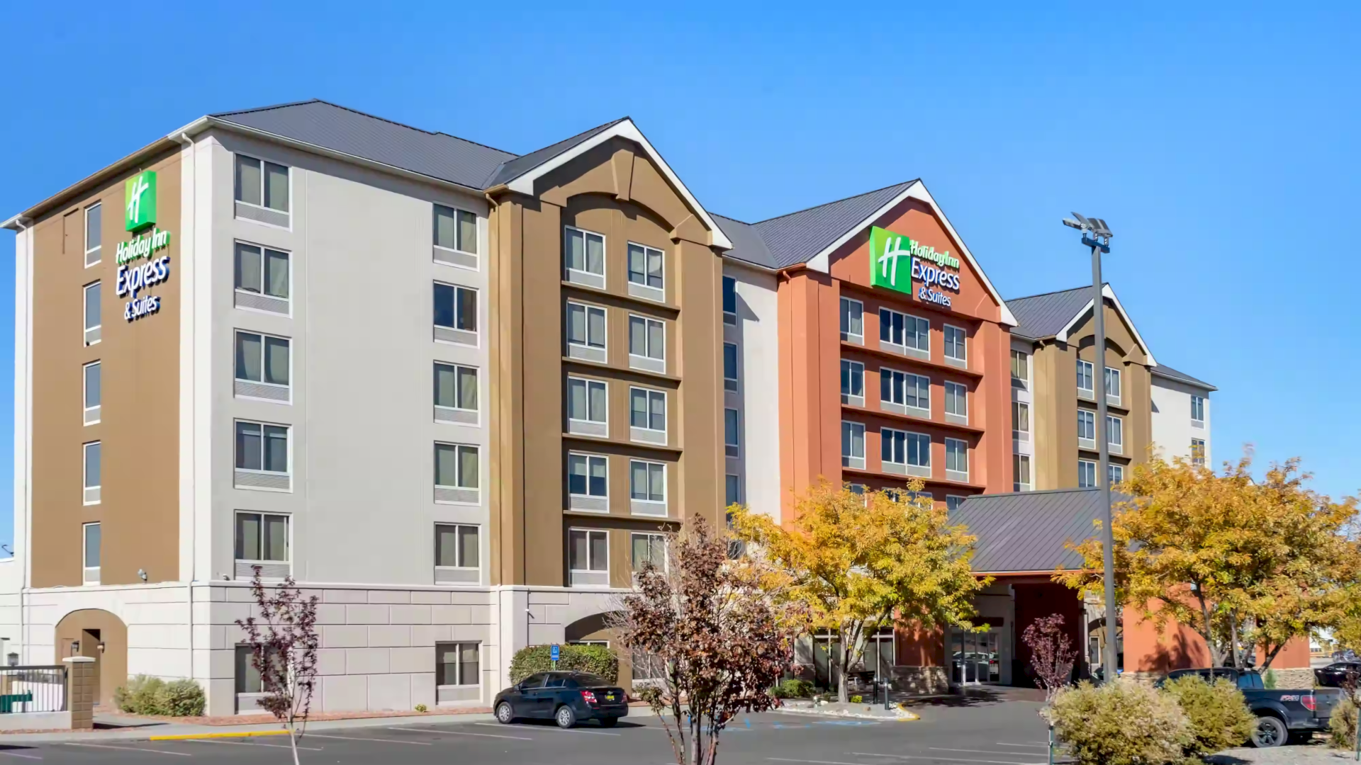 A multi-story hotel building with a modern design, surrounded by trees and a parking lot with cars, under a clear blue sky.
