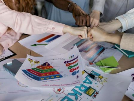 A group of people are fist bumping over a table with various colorful charts and documents, indicating teamwork and collaboration.