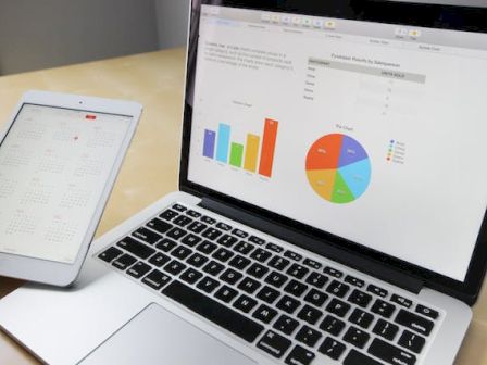 A laptop displays bar and pie charts while a person holds a tablet showing a calendar, both on a wooden desk.