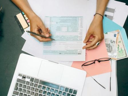 The image shows hands completing paperwork on a desk, surrounded by a laptop, glasses, folders, and various currency notes.