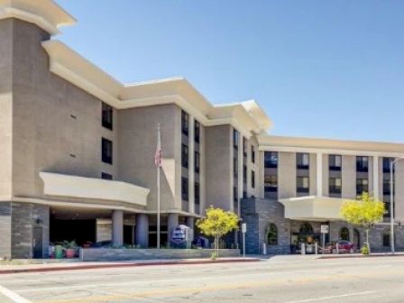 A multi-story building with modern architectural features, parking area, and trees in front, situated along a street with clear skies above.