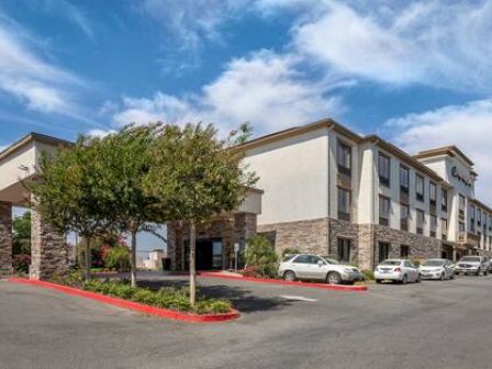 The image shows the exterior of a hotel with several parked cars, trees, and a partly cloudy sky.