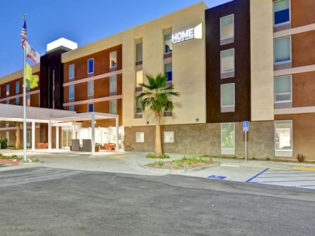 The image depicts the exterior of a modern hotel, displaying the sign "HOME." There are multiple floors, accessible parking spaces, and flags flying.