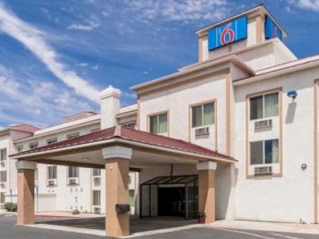 The image shows a three-story hotel building with a large, blue "6" logo on top, a covered entrance, and a clear sky above.